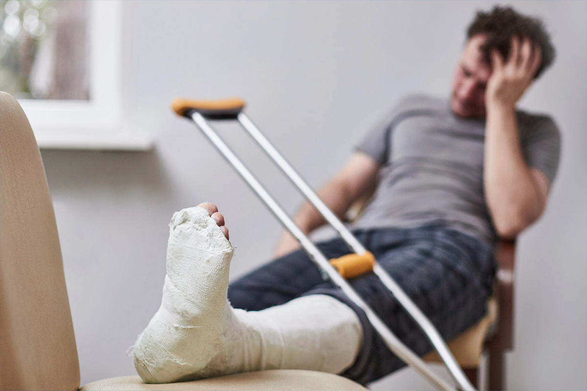 Man with a wrapped injured foot in a cast rests on a chair while crutches lean nearby, looking frustrated in a living room.