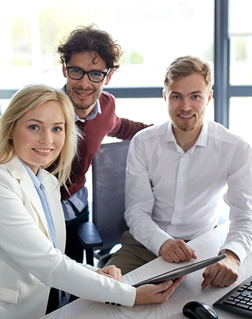 Woman in a white blazer hands a tablet to two men at a bright office desk with smiles on their faces.