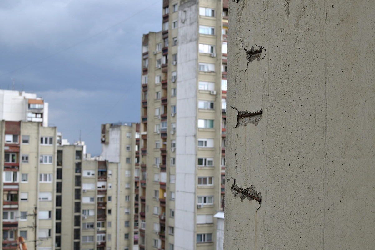 Close-up of a cracked concrete wall with rusted rebar in the foreground, urban high-rise apartments in the background.
