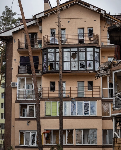 Beige apartment building with extensive damage, many broken windows and debris on balconies; leaning trees in front.