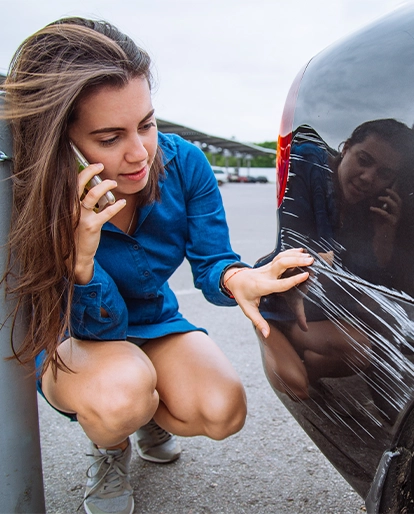 Woman crouching beside a dark car, talking on a phone while inspecting a scratch on the bumper