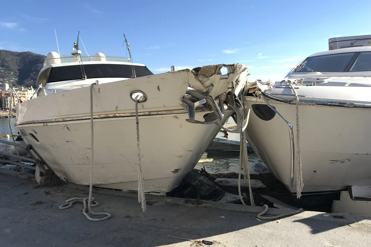 Two white motor yachts tied at a dock, with the left boat's bow heavily damaged from a collision and mooring ropes hanging around.