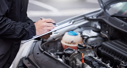A mechanic or technician wearing a dark jacket writes on a clipboard while inspecting the open car hood and engine bay.