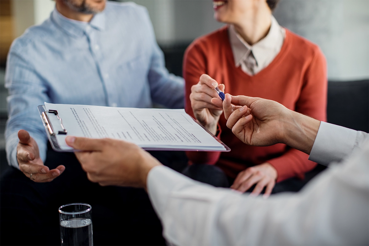 People in a business meeting reviewing a document on a clipboard; one person points with a pen while others listen.