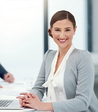 Smiling businesswoman in a light gray cardigan sits at a conference table with hands clasped, in a bright office.