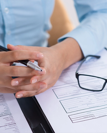Close-up of hands holding a pen over forms on a desk, with glasses nearby.